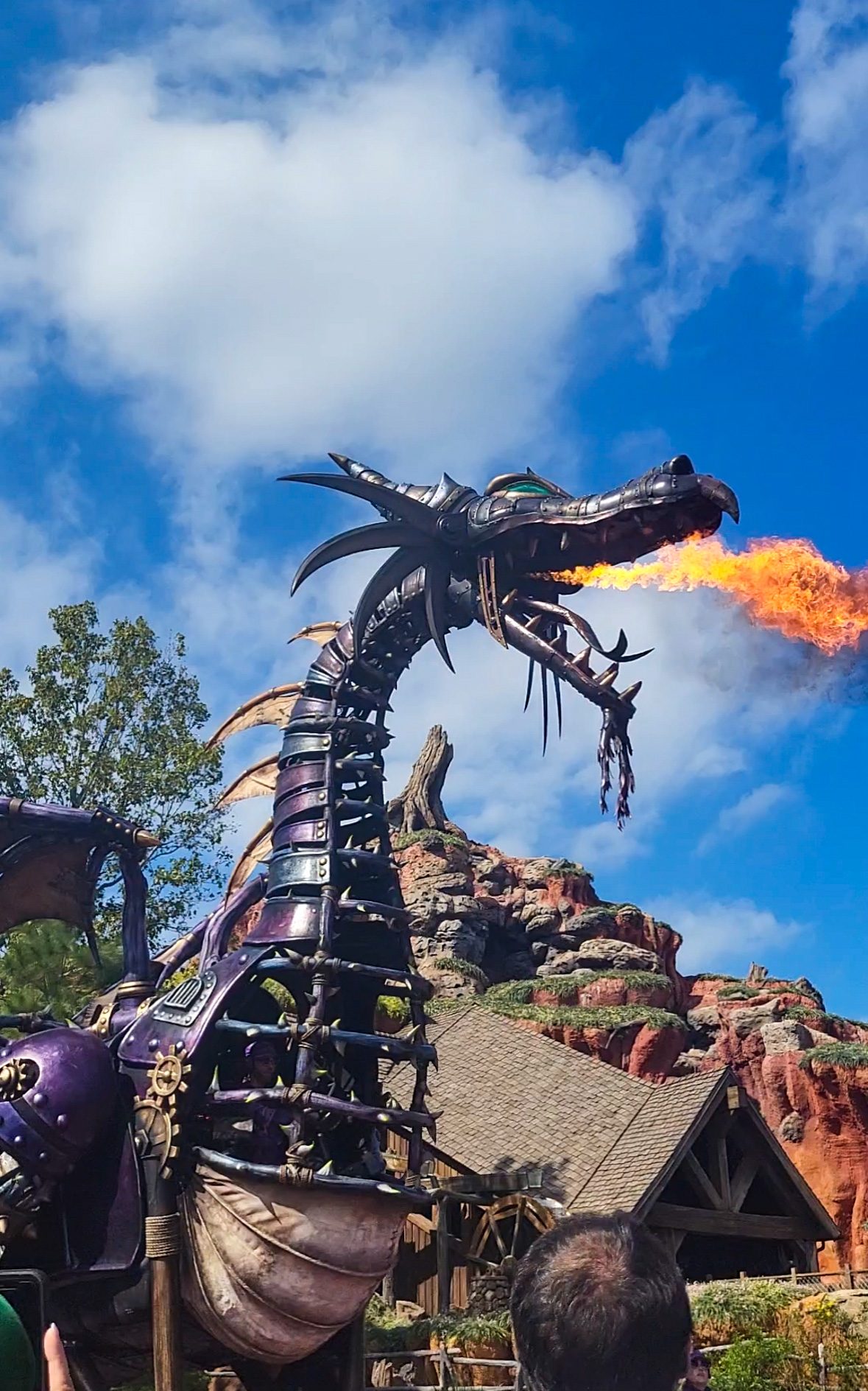 An image of a purple and black metal dragon breathing fire in front of blue skies and a brown and green man-made mountain. 