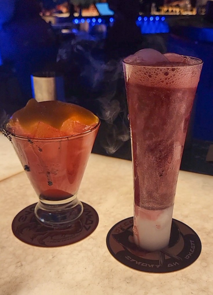 An image of two drinks in a low light bar. The left one is small, orange, has black drips down the glass, is full of ice, and has foam on top. The right one is tall, pink in colour with swirling glitter, and is bubbling. 