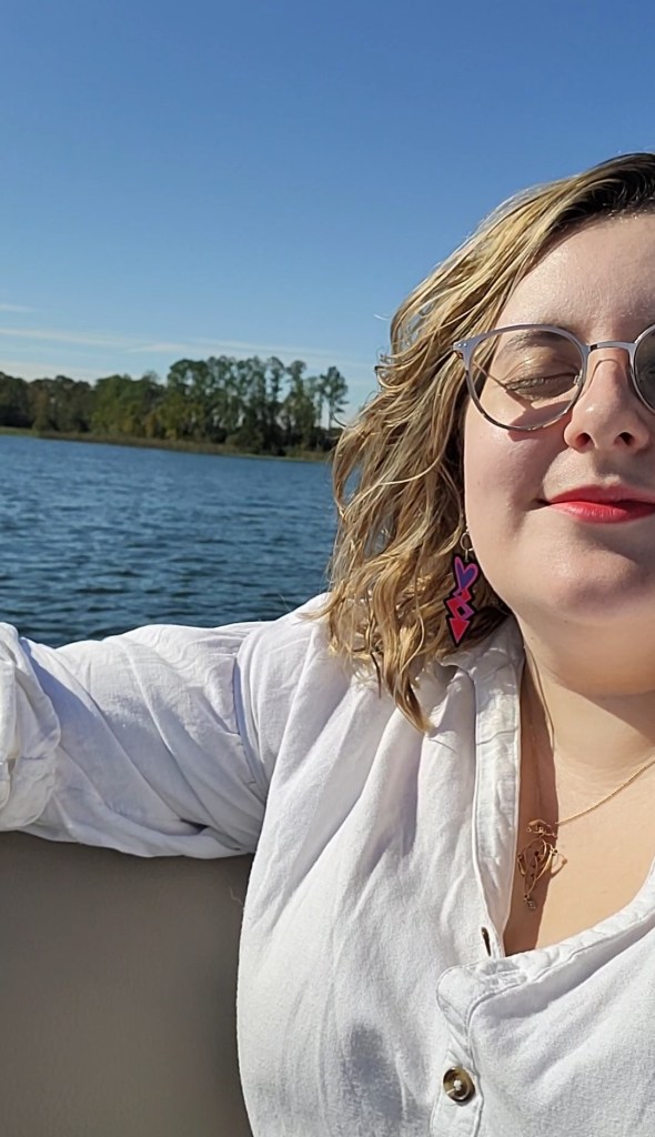An image of Rosie smiling into the sun in front of a blue lake. She is a white woman with a blonde wavy bob, glasses, and is wearing a red lip and a white blouse.