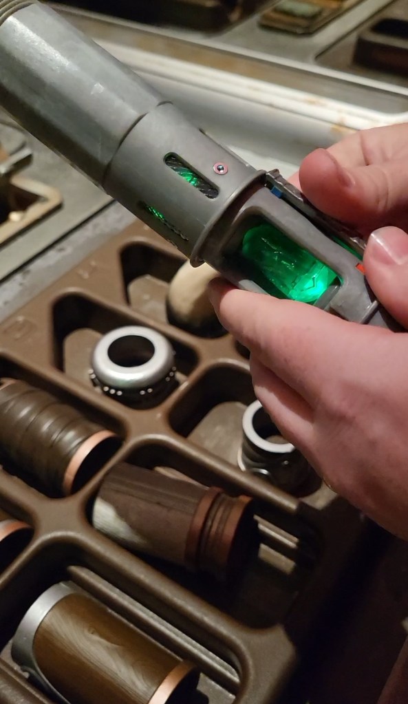 An image of a green kyber crystal being  slotted into a grey lightsaber. There are lightsaber parts in a brown tray below it. 
