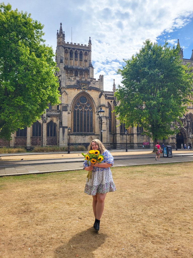 AN image of Rosie, a white woman with wavy blonde hair and glasses, holding a bunch of sunflowers. She is stood infront of a grey-yellow church by two green trees. She is wearing a blue and white mini dress and black boots. 