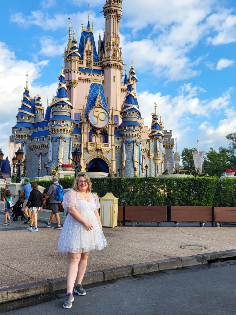 An image of Rosie, a white woman with short blonde wavy hair and glasses, stood infront of the pink and blue castle at Magic Kingdom. Rosie is wearing a white mini dress with .yellow flowers, grey sneakers, and a blue backpack. She is facing the camera. 