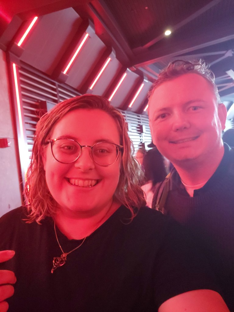 VelociCoaster queue An image of Rosie and Rory looking damp and lit by red lighting. They are both white. Rosie has shoulder blonde wavy hair and glasses. Rory has short light brown hair.
