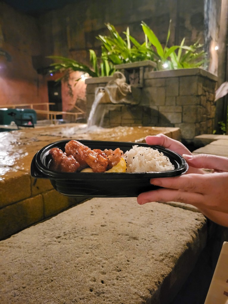 honey sesame chicken at Yak & Yeti An image of two white hands holding a black bowl of chicken, vegetables and rice. Behind the bowl is a fountain and greenery.