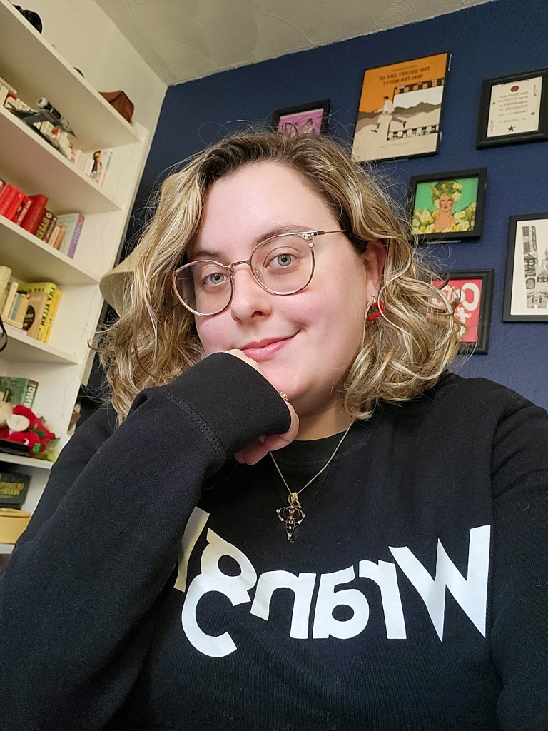 An image of Rosie, a white woman with curly blonde hair and glasses, looking at the camera. She is wearing a black crew neck jumper that says 'Wrangler' in white. 