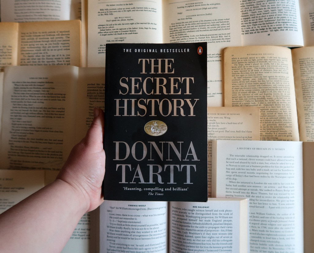 An image of a black cover book, that says 'The Secret History' and 'Donna' Tartt' in gold letters. It is being held by a white hand over yellow and white pages of books.
