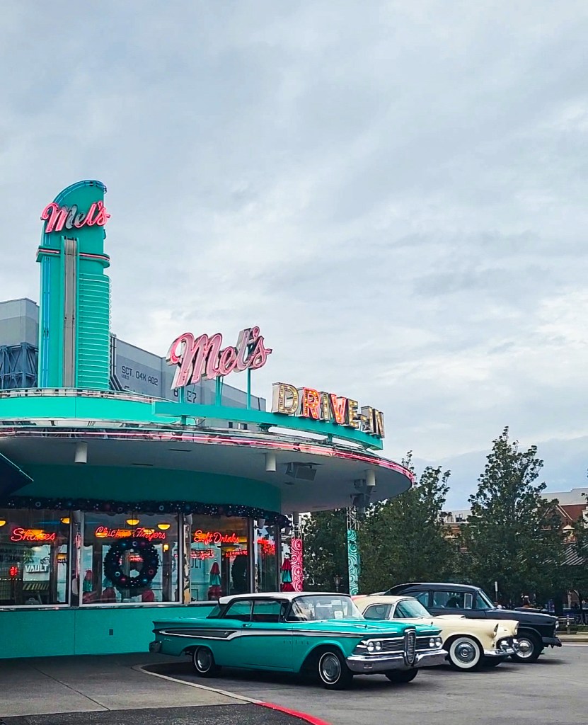 Mel's Drive In An image of the vintage inspired diner, Mel's Drive In. There are three classic cars out in front of the blue building. The building has neon signs with the name on it.