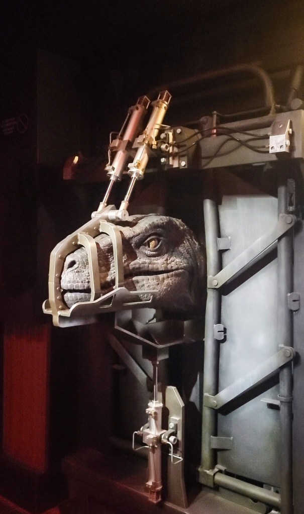 animatronic velociraptor An image of a grey animatronic velociraptor in a grey cage. It has yellow eyes and has a grey muzzle on.