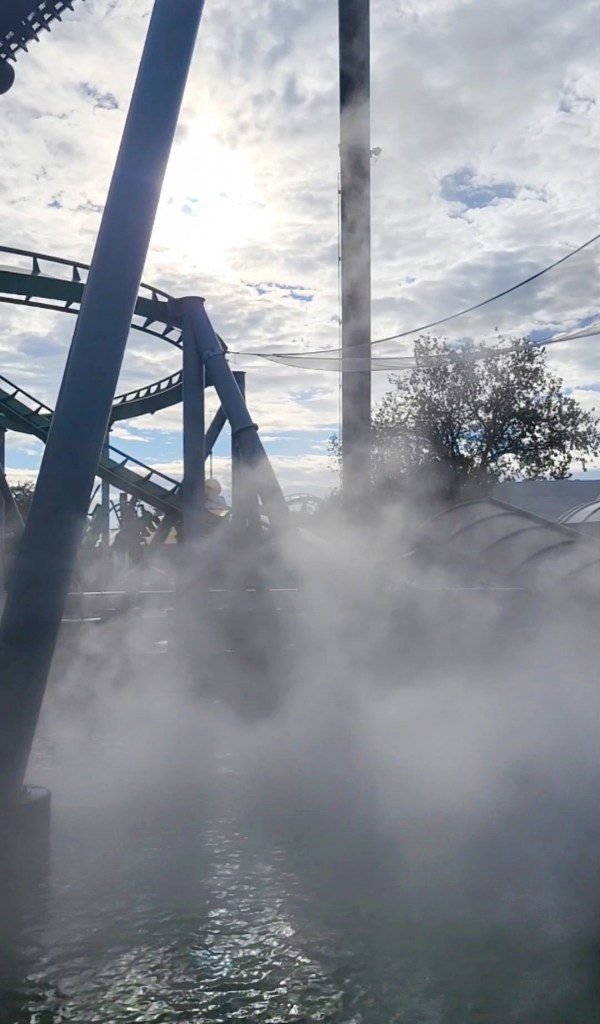 hulk coaster A sprawling track of a roller coaster covered in mist coming form a tunnel. The sky behind it is blue and cloudy.