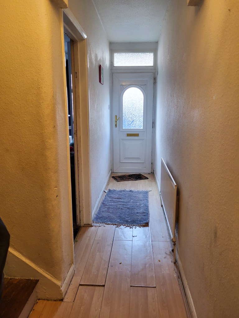 An image of a long hallway. There is a white door with a window in the middle and a window above. The walls are off-white and grotty, and the floors are laminate light wood and also grotty. There is a purple rug in the centre, and a radiator on the right wall. 