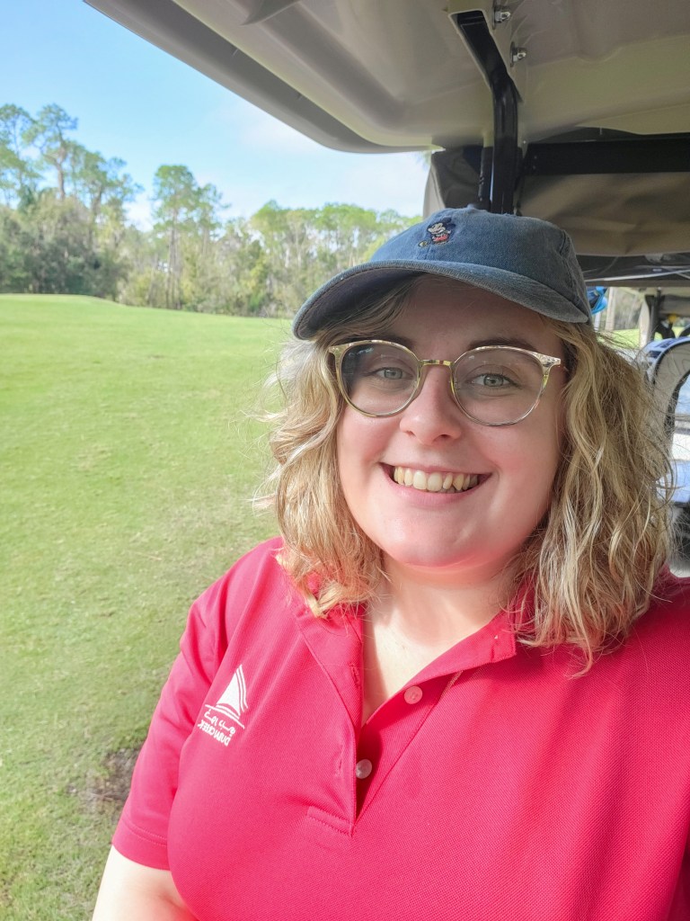 An image of Rosie, a white woman with short blonde hair and glasses, taking a selfie. She is in a golf cart, wearing a red polo, and a blue cap.