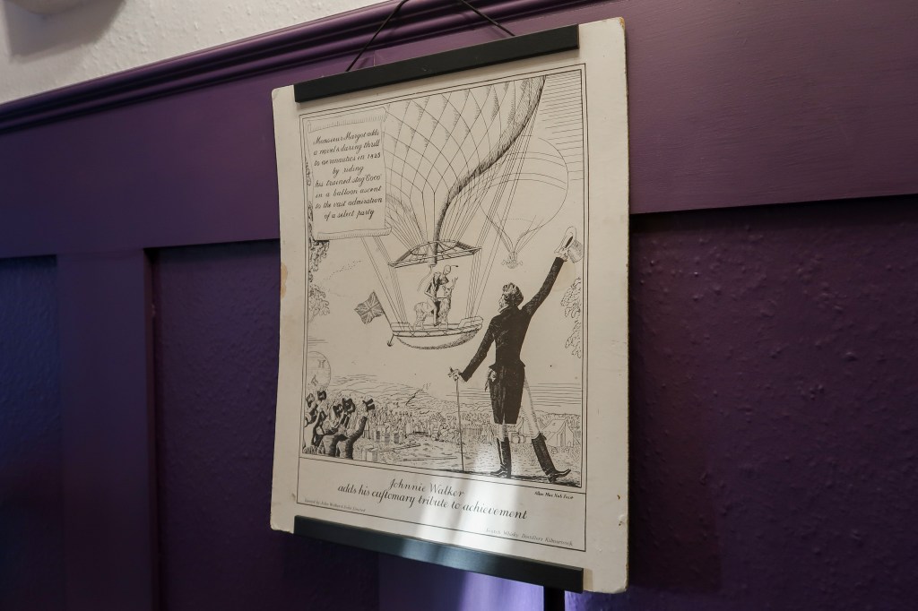 A Victorian print hanging up on the wall. The wall is purple and panelled. The print is a man waving a hot air balloon on, and is a Johnny Walker ad. 