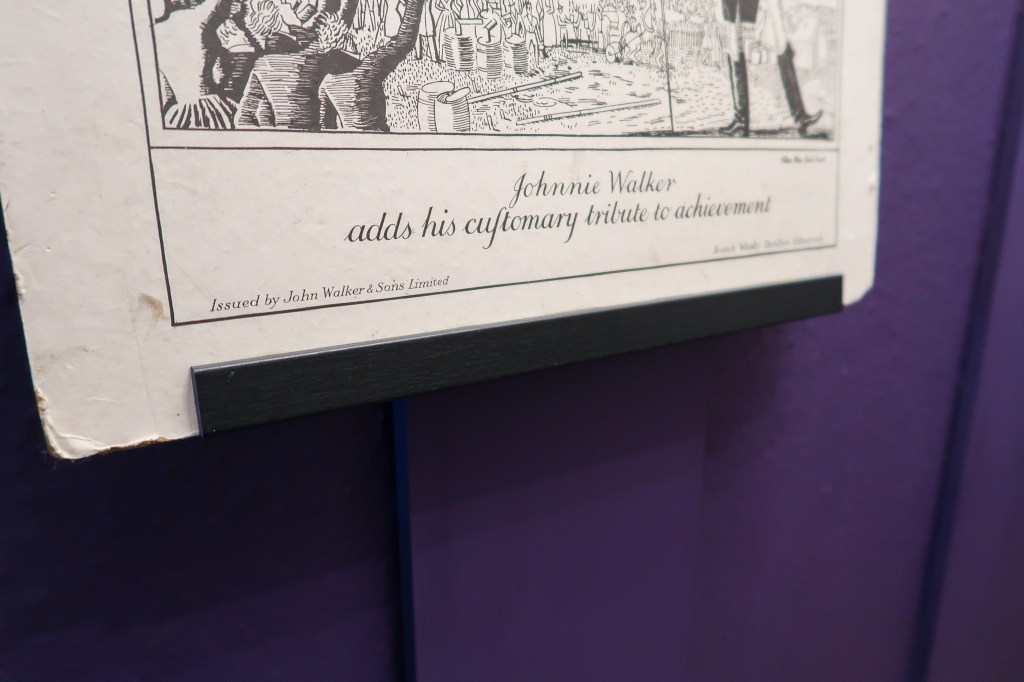 A Victorian print hanging up on the wall. The wall is purple and panelled. The print is a man waving a hot air balloon on, and is a Johnny Walker ad. 