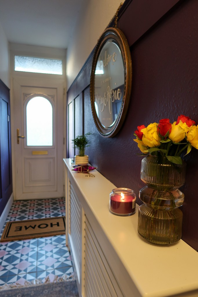 A shot of a hallway. There is a white door with a window in the middle and a window above at the end of the hallway. The walls are purple, the floors are pink and navy tiles, and there is a white radiator cover. On top of the cover are flowers, a candle, knickknacks, and a gilded mirror. 