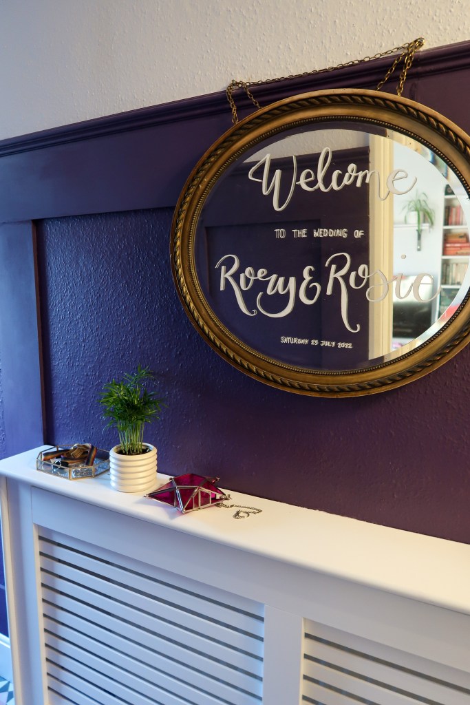 An image of a gold mirror hanging over a white radiator cover. The wall behind is purple and panelled. There are knickknacks and a plant on the radiator cover. 