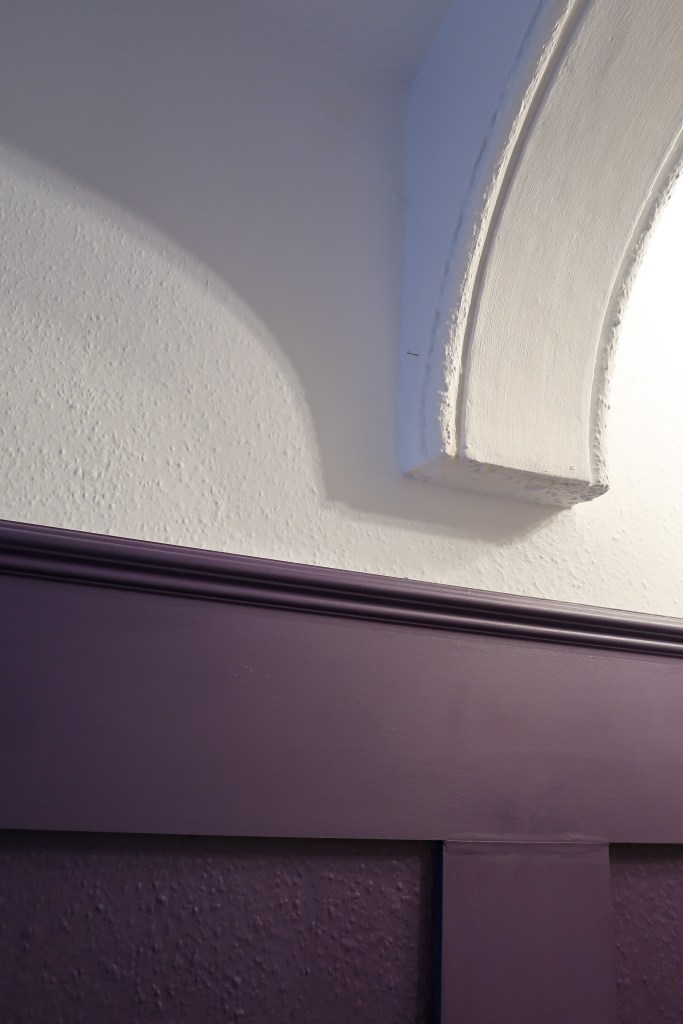 An image of our lovely painted walls! There is a white arch at the top of the wall, and the top of the wall is white. The bottom two thirds are purple panels, with a purple dado rail. 