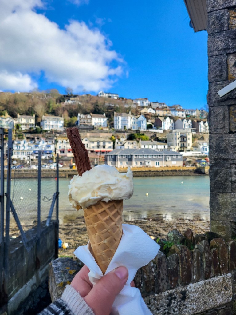 An image of a white ice cream in a cone with a flake, in front of the blue sea and cliffside houses. 