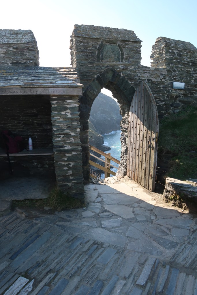 A grey stone wall and floor, with a doorway. The doorway leads out the roaring blue sea and a wooden staircase.