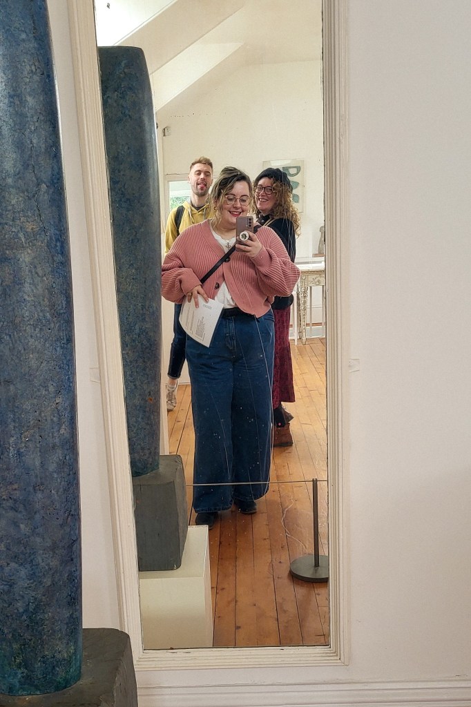 An image of Rosie, a white woman with dyed blonde hair and glasses, taking a selfie in a tall mirror. She is wearing blue wash jeans and a pink cardigan. Her mum and brother are behind her.