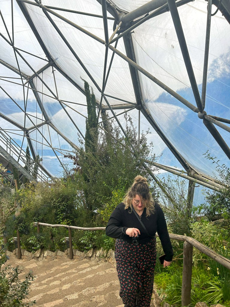 An image of Rosie in a black dress, walking up the stairs at the Eden Project. She is surrounded by green plants and is looking at her camera. 