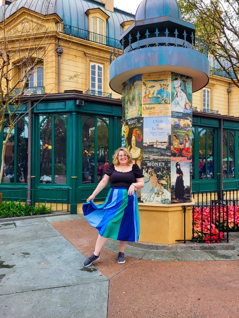 An image of Rosie (a white woman with curly blonde hair and glasses), stood in front of a pillar that is covered with art posters. She is wearing a black bardot top, and a blue and green striped skirt. She is holding the skirt in her right hand. The walls and windows around her are yellow and green.