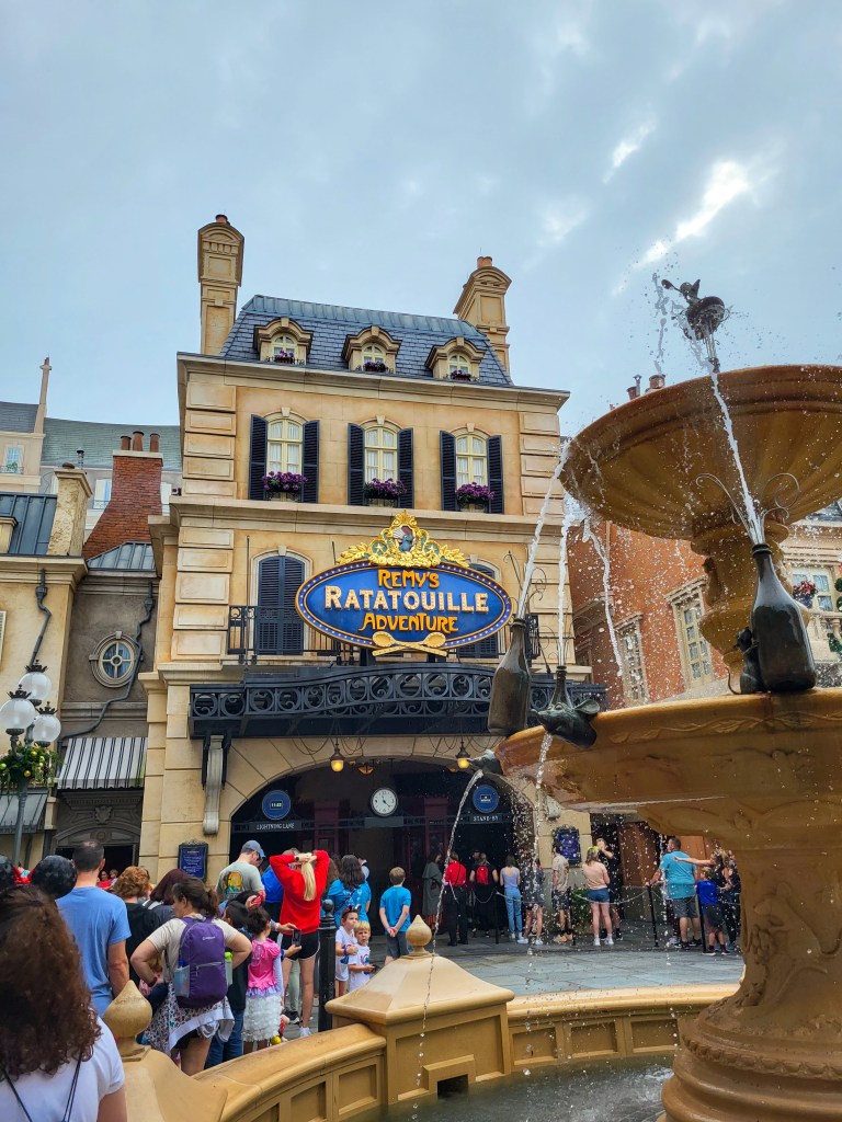 The front entrance to Remy's Ratatouille Adventure. The building is yellow stone and there is a blue sign with the ride name on front. There is a yellow stone fountain with bottles and metal rats spouting water.