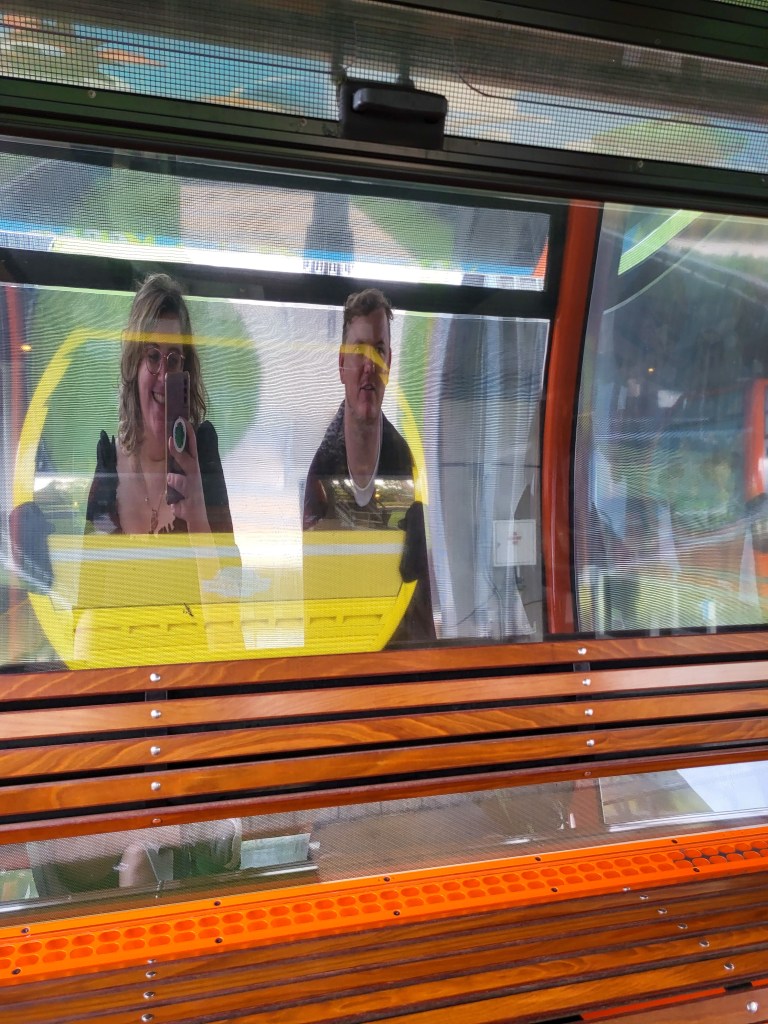A photo of Rosie taking a selfie of her and Rory in the window of the Skyliner. They are elongated like a funfair mirror. There is a wooden bench in front of then