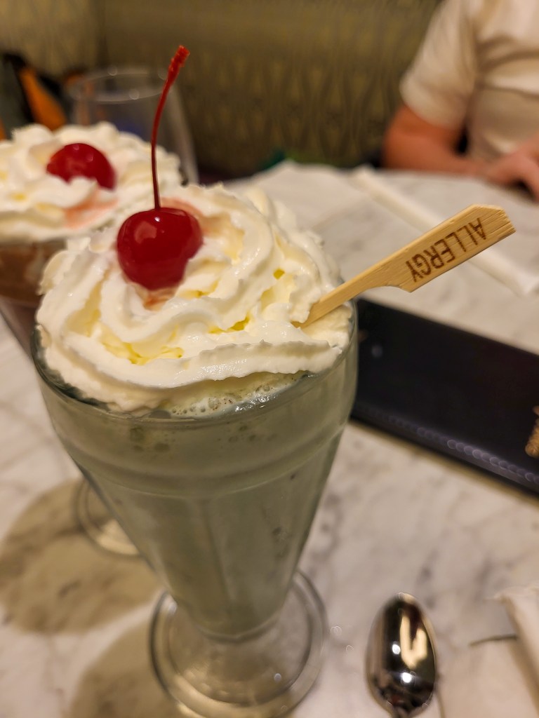 A light green, mint choc chip milkshake in a clear glass with whipped cream and a red cherry on top. There is a wooden stick in the cream saying 'allergy'.