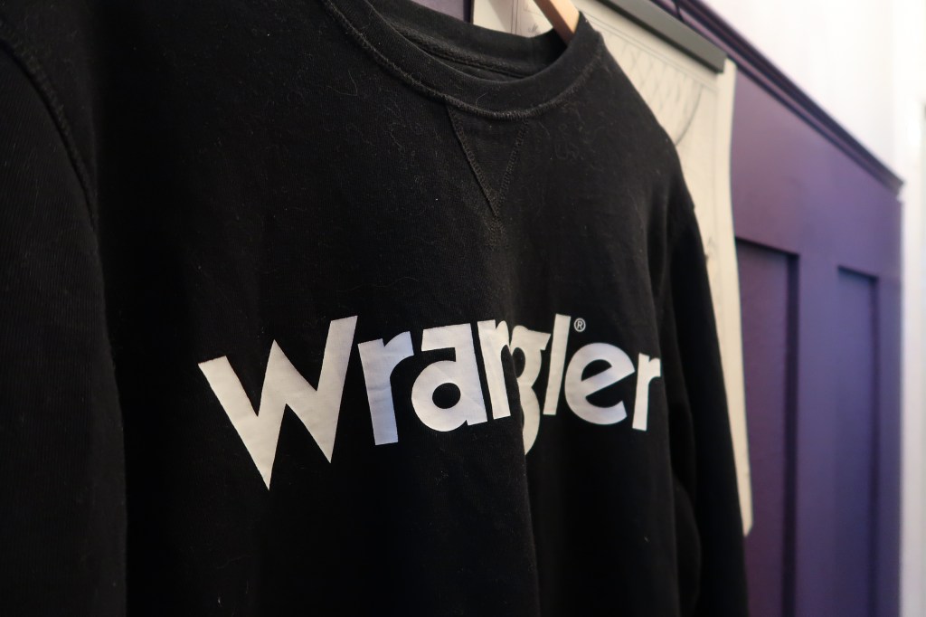 A black sweatshirt reading 'Wrangler' in white writing, against a purple wall