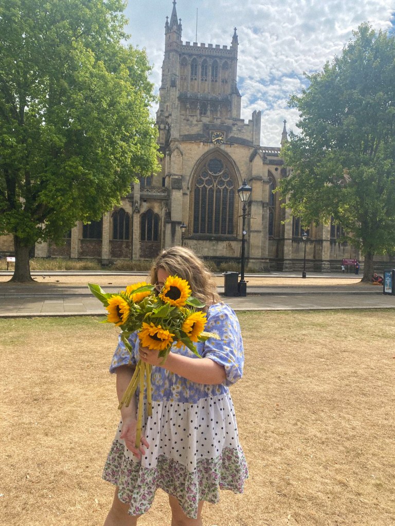 An image of Rosie, a white woman with wavy bobbed blonde hair, in front of Bristol Cathedral. She is holding a bouquet of sunflowers and is wearing a mini smock dress. The dress is light blue at the top with white flowers; white with pink flowers at the bottom; and white with black dots in the middle.