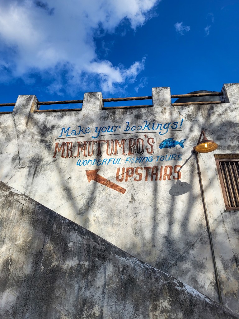 Blue skies above a white, weather worn building. The building reads in red and blue "make your bookings! Mr Mufumbo's wonderful fishing tours upstairs"