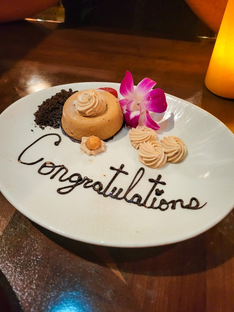 A white oval plate topped with chocolate crumb; a glittery gold caramel mousse with swirls on top; a pink flower; more swirls; and the word 'congratulations' written in chocolate