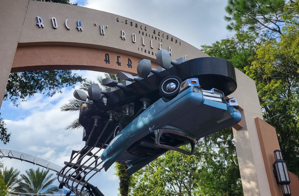 An image of a classic light blue car, hanging upside down at the entrance to Rock n Roller Coaster. It is sat on the head of an electric guitar.