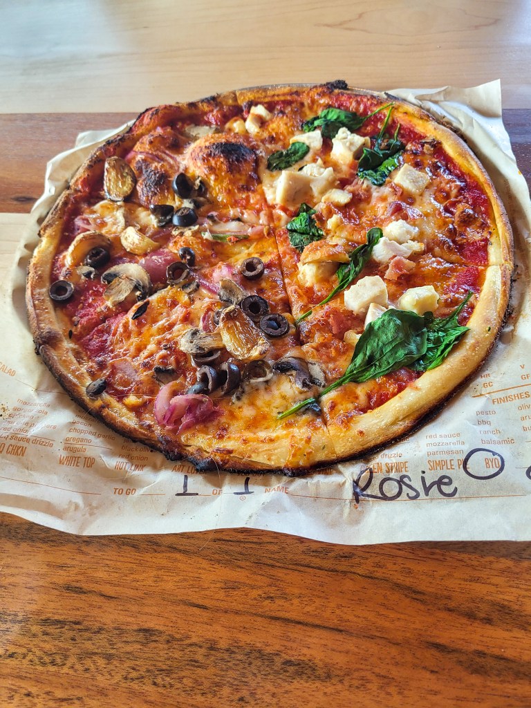 A stonebaked pizza with tomato sauce and cheese. On the left side it is covered in onion, mushrooms, and olives. The right side is covered in basil and chicken. It sits on a brown piece of paper that says 'Rosie' in black