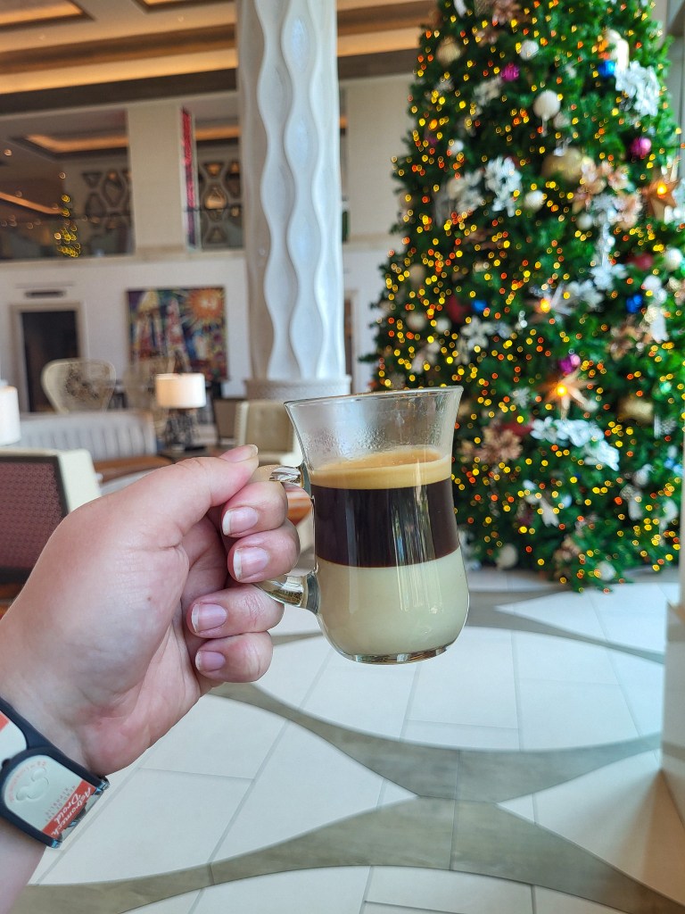 A white woman's hand holding a clear coffee mug. There is condensed milk in the bottom, and espresso on top. Behind the drink is a green and gold Christmas tree and white pillars