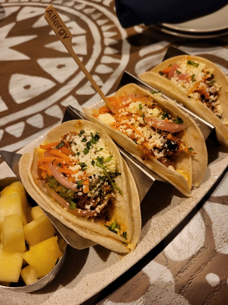 Three double-stacked tacos in a silver holder. They are all full of meat, carrots, slaw, and topped with cheese. The middle one has a brown stick that says "allergy" on it. To the left is a silver bowl of chopped pineapple. This all sits on a mosaic style table in brown and white