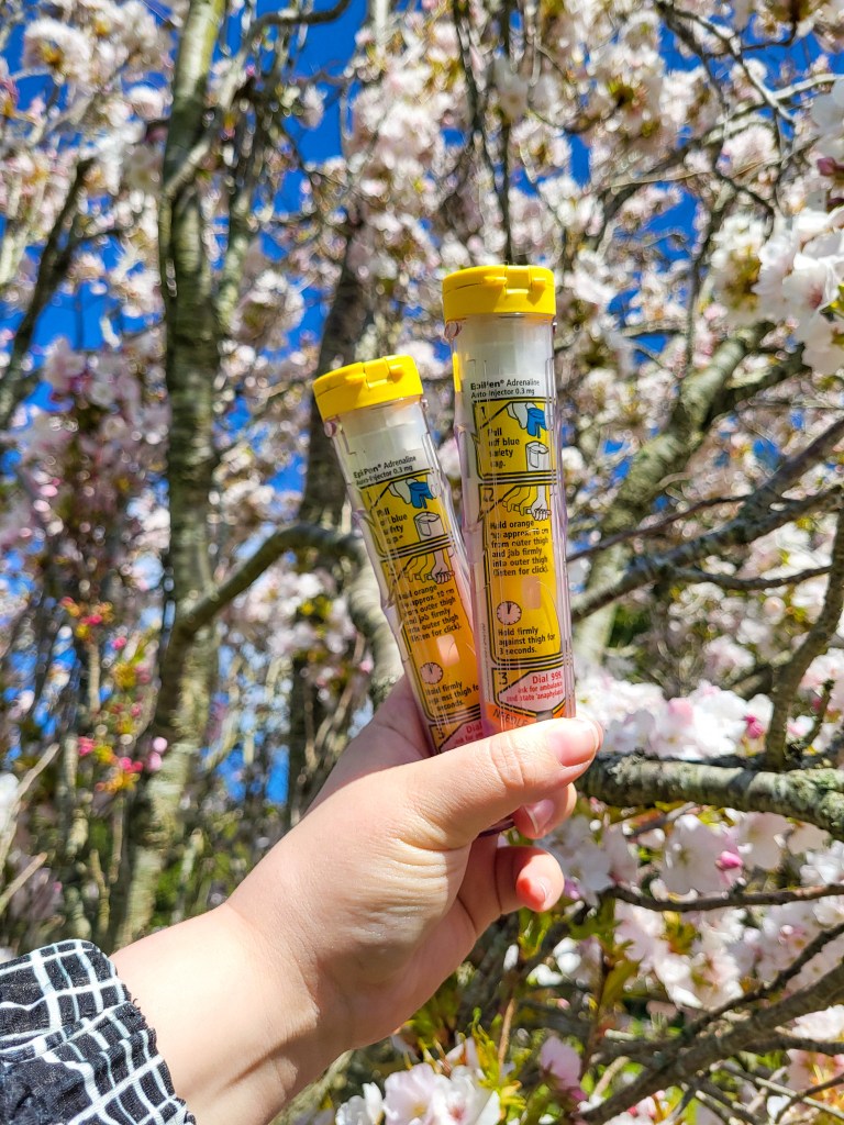 An image of a white hand holding two EpiPens - white needle cartridges with yellow lids and yellow boxes. They are being held in front of pink and white flowers against a blue sky 