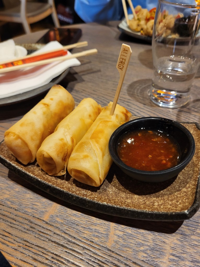Three golden spring rolls on a brown speckle, rectangle plate, with a chilli red dipping sauce to the right in a black ramekin. There is a brown stick that reads 'allergy' on the third roll. 