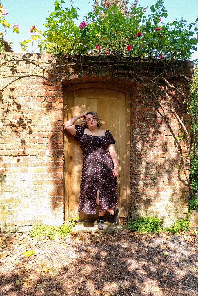 An image of a Rosie (a white woman, with a curly blonde bob, and glasses) standing within an archway and in front of a brown door. She has her right arm leaning against the arch and her hand against her head. Her other hand is down by her side. She is wearing a black midi dress with puff sleeves and a cherry print and is smizing at the camera. Around the door is red brickwork with the leaves, flowers, and branches of a rose bush
