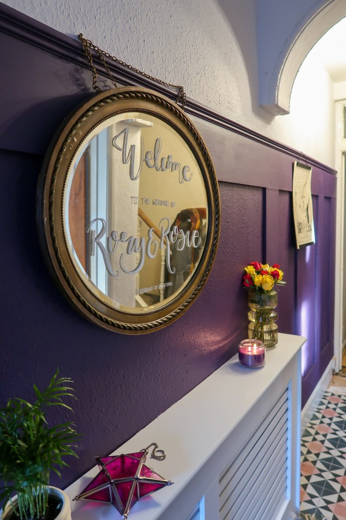 A picture of a hallway. A purple panelled wall runs along tje left, with a white ceiling above it. There is a white arch towards the top right of the image. The centre of the image focuses on a gold mirror above a white radiator cover. The gold mirror says 'welcome to the wedding of Rory & Rosie' in white cursive. On top of the radiator cover is a green plant, a pink glass star, a purple candle, and a bunch of flowers. The floor peeks from the bottom of the image; pink, white, and blue tiles. 