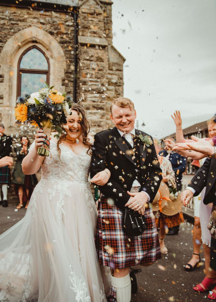 An image of Rory and Rosie leaving a grey/brown church, with onlookers showering them in confetti. On the left, Rosie is a white woman with long brown hair in waves, in a white and lace wedding dress, and is holding a yellow and green bouquet in her right hand. Her left hand is linked through Rory's right arm. He is on the right of the picture. Rory is a white man with light brown hair, and is wearing a black Prince Charlie jacket, with a red and blue kilt, black sporran, and long white socks. The couple are grinning. 
