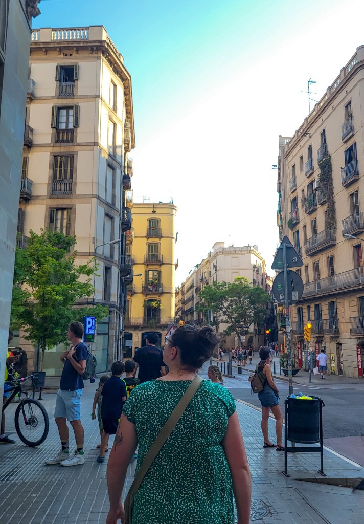 A woman with brown hair in a bun and wearing a green dress looks up at the Gothic Quarter skyline to the left. In front of her are a group of people. In front of them are yellow and sandstone coloured buildings with balconies. 