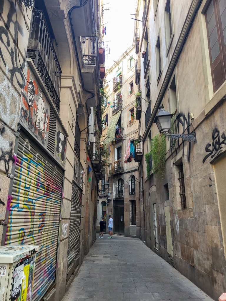 A side street in the Gothic Quarter, that forks to the left. The sandstone-esque blocks of the bottom half of the buildings have shutters and graffiti on them. The windows on the walls have blankets or curtains hanging. 