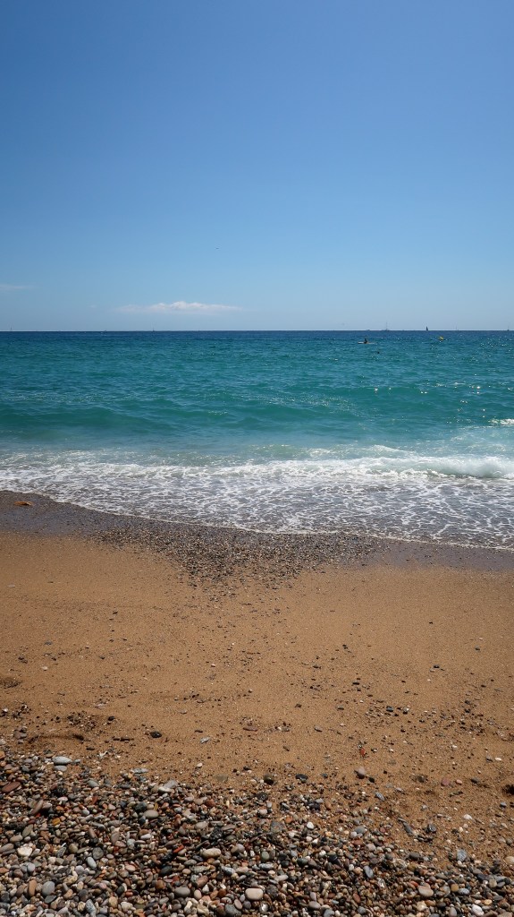 A clear blue sky leading down to a beautiful blue ocean. It is slightly choppy but the sea foam laps onto golden sand. At the seashore, there are rocks scattered across the sand. 
