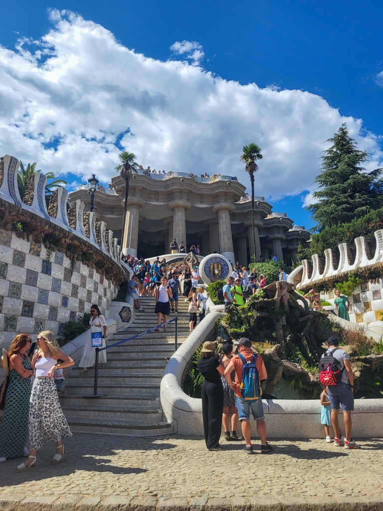 A white pillared building  with curved steps leading down the front. The walls on the side are chequered and there is a green, foliage covered statue at the front. Crowds of people stand on the stairs. 