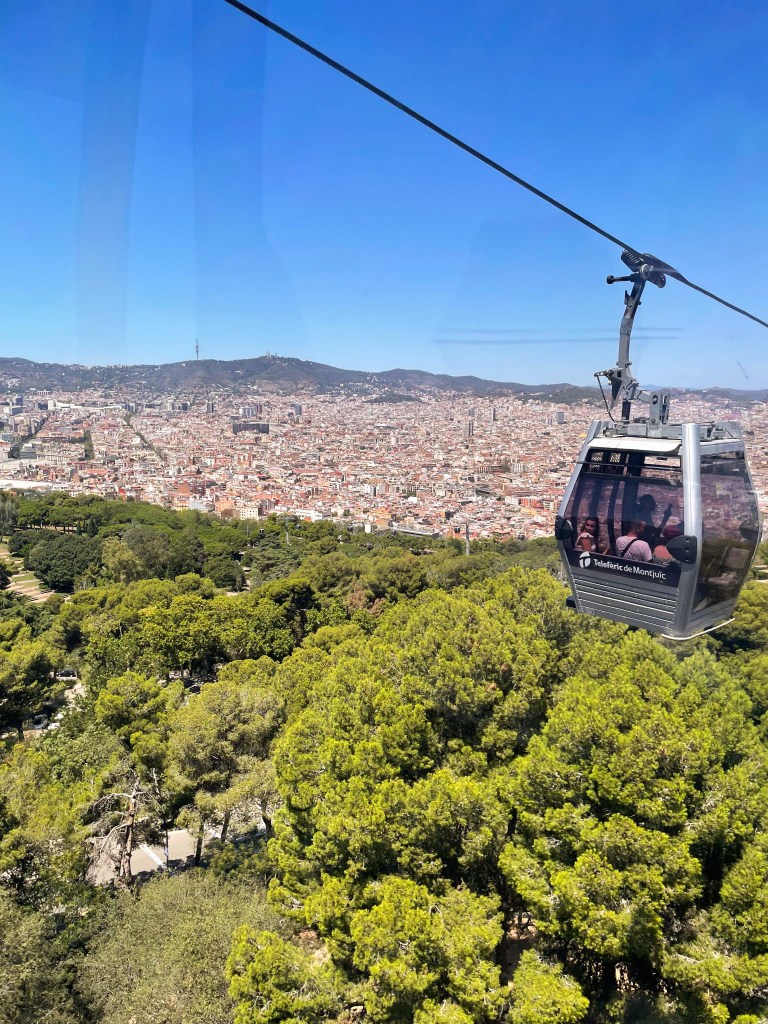 A view from a cable car on the way down from Montjuïc. Green trees fill the bottom of the image, and the orange city blocks of Barcelona become clear in the middle. Above them are dark hills and a clear blue sky. A cable and a cable car sit on the right of the image. 