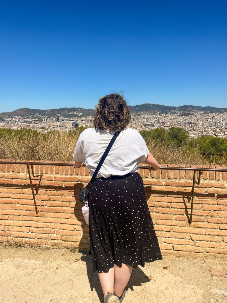 Rosie, a white woman with a blonde and brown bob, stood with her back to the camera. She is holding on to a railing and looking out at the view. She is wearing a white t-shirt and a black wrap skirt with white dots. There is a black bag strap across her back, leading to a small black bag and a pink waterbottle. The view behind her is of blue skies, dark green hills, and the city of Barcelona. 