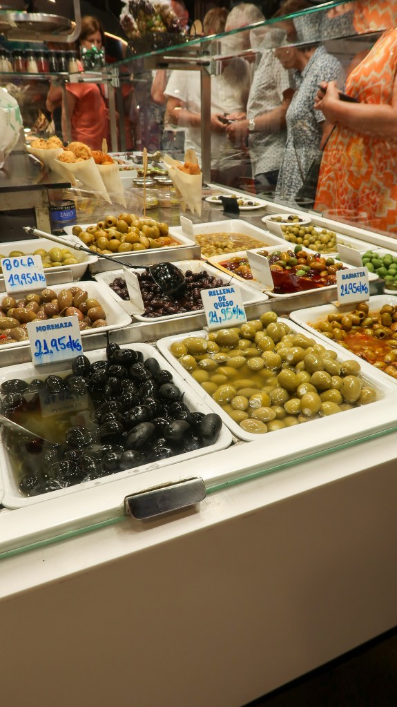 White trays filled with varieties of green and black olives behind a glass screen. Behind them are spoons and cones of crispy food. There are crowds of people towards the back of the image behind glass. 