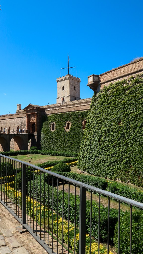 A pale sandstone coloured fortress covered in green foliage, with a blue sky behind it. There is a bridge on the left to the entrance, and a black railing at the bottom of the image. 