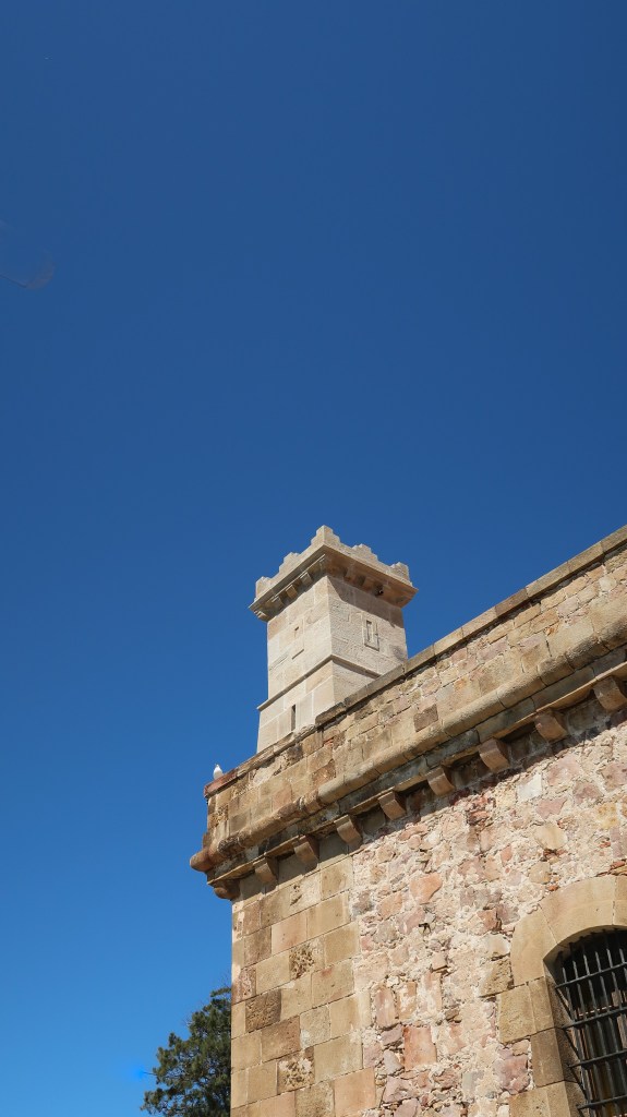 A clear blue sky pierced by a sandstone coloured tower and wall of the fortress. 